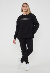 Set di felpa e pantaloni neri con design oversize; la felpa presenta un logo EA7 testurizzato. Indossato con sneakers bianche.