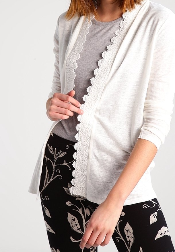 Cardigan blanc avec bordure en dentelle, porté sur un haut gris, complété par des leggings à motifs floraux noirs. Tissu doux et léger.
