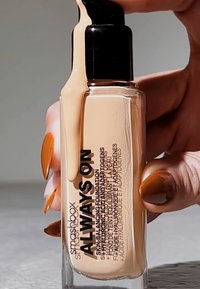 Fond de teint liquide beige dans un flacon en verre transparent avec une pompe noire. Les étiquettes présentent un texte en gras noir. Consistance moyenne. Main avec du vernis à ongles orange.