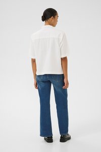 Chemise blanche à manches courtes avec un dos en yoke, assortie à un jean droit bleu et des chaussures noires, vue de derrière.