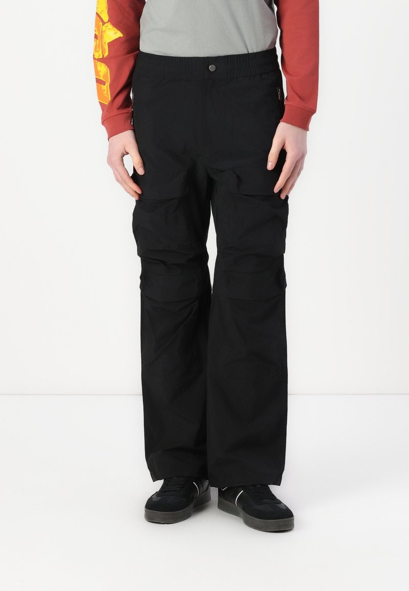 Personne portant un pantalon cargo noir avec des poches latérales, des baskets noires et un t-shirt rouge à manches longues avec un motif jaune sur la manche.