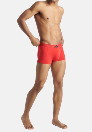 Boxer aderenti rossi realizzati in tessuto elasticizzato con un cinturino nero che presenta un logo. Il design è aderente e di lunghezza a metà coscia.
