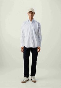 Camisa branca de botão de mangas longas, calças azul-marinho, boné bege e ténis brancos com detalhes em bege, em pé sobre um fundo claro.