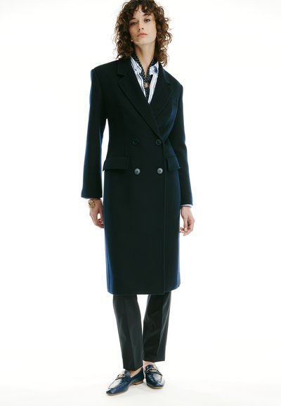 Femme aux cheveux bouclés portant un long manteau noir croisé, une chemise rayée, un pantalon noir et des mocassins foncés avec détails à boucle dorée.