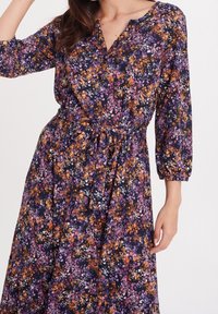 Žena v dlouhých rukávech s páskovými šaty, které mají malý květinový vzor v purpurové, oranžové a bílé barvě na tmavém pozadí.