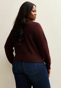 Kobieta z długimi ciemnymi włosami, ubrana w burgundowy sweter i ciemnogranatowe dżinsy, lekko odwrócona w bok na tle jasnym.