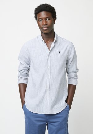 Joven con cabello rizado y oscuro, vestido con una camisa de botones a rayas azul claro y pantalones azules, de pie con las manos en los bolsillos contra un fondo blanco.