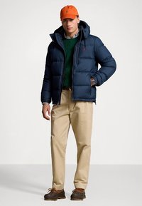 Mornarsko moder puhasti jakna s kapuco, zelen pulover iz pletenine, bež hlače in rjavi škornji. Z oranžno kapo in srajco z vzorcem pod njim.