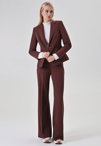 Bordeaux op maat gemaakt pak met een fitted blazer met een ronde gouden knoop, gecombineerd met een crèmekleurige structuurtrui en een brede broek. Witte sneakers.