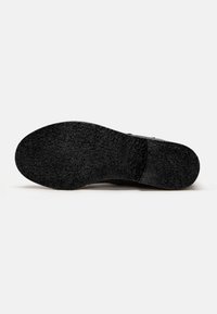 Schwarze Schuhsohle aus strukturiertem Gummi, mit flachem Design und leichtem Absatz. Eingraviertes Markenlogo in der Nähe des Absatzbereichs sichtbar.