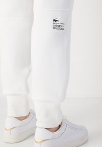 Vita sweatpants med ribbad mudd, prydda med en Lacoste-logotyp. Parat med vita sneakers med detaljer i guld och beige. Slät textur.
