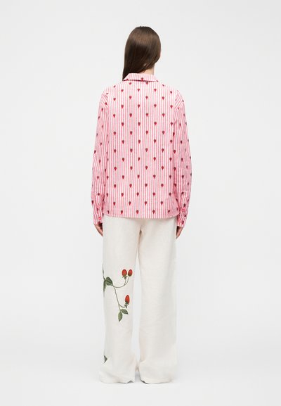 Woman with long brown hair wearing pink and white striped shirt with red ice cream cone patterns and white pants with strawberry embroidery, standing.