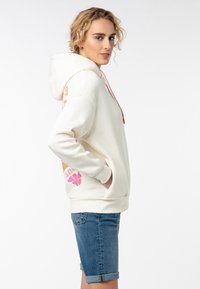 Crème hoodie met roze trekkoorden en grote grafische details in roze en oranje. Heeft een voorzak en ribgebreide manchetten. Casual fit.