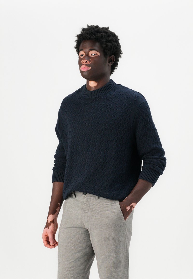 Pull en maille marine avec un motif texturé et un col en rib, associé à un pantalon gris clair à motifs, créant une tenue complémentaire.