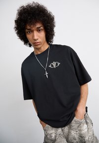 Black oversized t-shirt with a silver graphic design on the chest. Paired with light gray patterned shorts and silver chain necklace.
