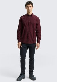Chemise à manches longues à carreaux bordeaux, avec boutons sur le devant, col, portée avec un pantalon sombre et des chaussures sombres, se tenant contre un fond clair.