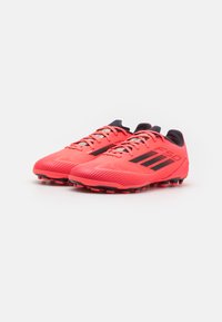 Botas de fútbol de color rojo brillante con una textura estriada, acentos negros y un diseño de perfil bajo. Presentan rayas y cordones negros en contraste.
