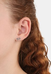 Orecchini a perno argento, a forma di piccolo fiocco, con una finitura lucida e dettagli delicati, indossati all'orecchio di un modello.
