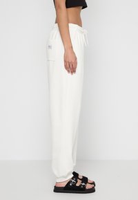 Cremfarbene Sweatpants mit elastischem Bund und seitlicher Tasche, aus weichem Material. Kombiniert mit schwarzen Sandalen mit Schnallenakzenten.