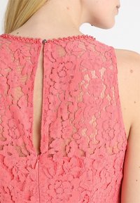 abito in pizzo corallo con motivo floreale, chiusura con bottone al collo e design con apertura a chiave sul retro. Tessuto morbido con sovrapposizione trasparente.