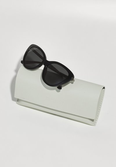 Black cat-eye sunglasses resting on a textured white leather case, featuring engraved branding and a sleek, minimalist design.