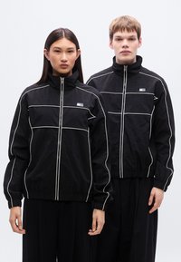 Μαύρα μπουφάν με φερμουάρ και λευκή περίγραμμα, με ψηλούς γιακάδες και ελαστικά μανσέτες, φορεμένα πάνω από μαύρο παντελόνι. Δύο μοντέλα στέκονται δίπλα δίπλα.
