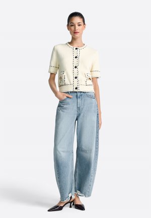 Knitted cream cardigan with black stitching details, short sleeves, and two front pockets, paired with high-waisted light blue denim jeans.