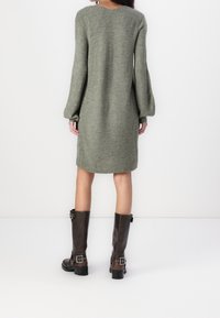 Robe en tricot vert olive avec des manches longues en forme de cloche et un ourlet côtelé. Associée à des bottes en cuir marron jusqu'aux genoux avec des boucles.