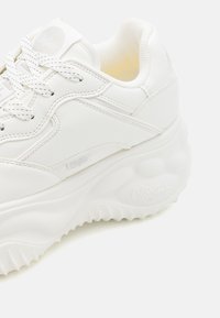 Zapatilla blanca con parte superior de cuero suave, silueta redondeada, suela texturizada y un branding minimalista en el lateral. Presenta cordones y costuras a tono.