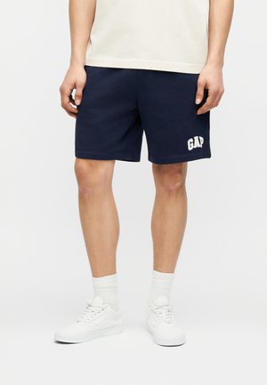 Uomo che indossa pantaloncini blu navy con il logo "GAP" sulla gamba sinistra, calzini bianchi a coste, sneaker bianche e una maglietta chiara.