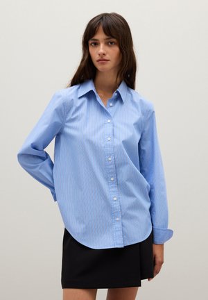Donna che indossa una camicia azzurra chiara a maniche lunghe con bottoni e strisce sottili, abbinata a una mini gonna nera, in piedi davanti a uno sfondo semplice.