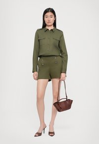 Olivovo-zelený outfit pozostávajúci z košele s golierom a vreckami na gombíky a šortiek s dvojitými gombíkovými akcentmi, doplnený malou hnedou kabelkou a topánkami na podpätku.