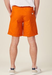 Shorts di cotone arancioni con orli arrotolati, tasche laterali e una tasca posteriore con un piccolo logo del marchio. Indossati con sneakers bianche e beige.