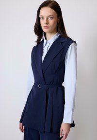 Navy blue tailored vest with a cinched waist, sharp lapels, and pleated design, layered over a light blue collared shirt.