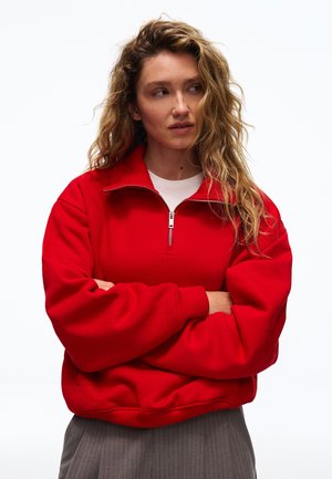 Jeune femme aux longs cheveux ondulés portant un sweat rouge zippé par-dessus une chemise blanche, bras croisés, regardant vers sa gauche.