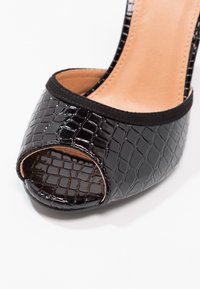 Chaussure ouverte en cuir verni noir avec un motif texturé de crocodile, dotée d'un intérieur beige lisse et d'une bordure noire autour de l'ouverture.