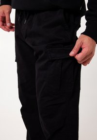Zwarte cargobroek van stevig materiaal, voorzien van grote zijzakken, een ontspannen pasvorm en een verstelbare tailleband met trekkoord.