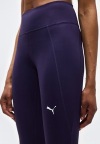 Κοντινό πλάνο ενός ατόμου που φοράει σκούρα μωβ κολάν Puma με εμφανείς ραφές και λογότυπο στο μηρό, τα χέρια χαλαρά δίπλα στο σώμα.