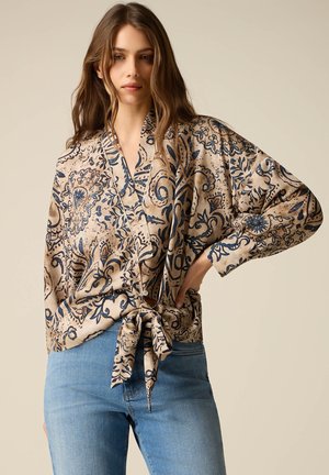 Vrouw draagt een beige blouse met lange mouwen en blauwe bloemenpatronen, gestrikt in de taille, gecombineerd met een lichtblauwe spijkerbroek, staand tegen een effen achtergrond.