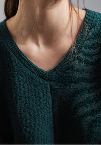 Maglione di colore teal, strutturato, con scollo a V. Vista ravvicinata che mette in risalto il motivo a coste del tessuto e la finitura morbida.