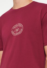 Burgunderfarget t-skjorte laget av myk bomull, med et sirkulært logo-trykk i lys beige plassert på brystet, og en klassisk rund hals-design.