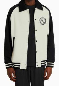 Vit och svart varsityjacka med tryckknappar, kontrasterande ärmar, en broderad logotyp och randiga muddarna. Mjuk tygtextur.