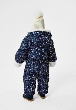 Combinaison de neige matelassée bleu marine avec motifs de fleurs blanches, col en peluche blanche et un bonnet assorti en tricot crème avec pompon. Chaussures robustes incluses.