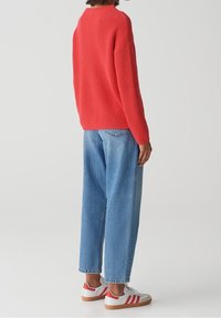 Person, der einen roten Strickpullover, lockere hellblaue Jeans und weiße Sneakers mit roten Streifen trägt und vor einem einfarbigen hellen Hintergrund steht.