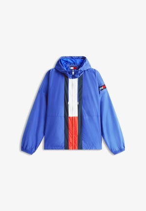 Cortavientos azul con capucha, con franjas verticales blancas, rojas y azul marino en el centro y un pequeño parche con una bandera en la manga izquierda.