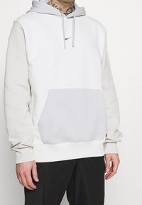 Sudadera con capucha en gris claro y blanco, con bolsillo tipo canguro, capucha ajustable con cordón y puños acanalados. Tejido suave con diseño en contraste.