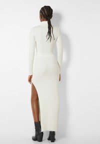 Femme aux cheveux tressés portant une robe blanche à col roulé côtelée avec une fente haute jusqu'à la cuisse, associée à des bottines à talons noires, vue de dos.