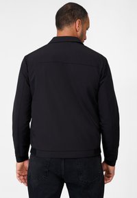 Zwarte lichte jasje met een kraag, gladde textuur en elastische manchetten, gecombineerd met een donkere spijkerbroek, gezien vanuit de achterkant.