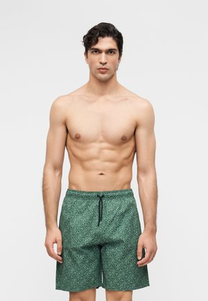 Homme musclé aux cheveux noirs ondulés, portant un short de bain vert à motifs avec un cordon noir, debout devant un fond clair uni.