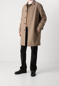Brauner Hahnentrittmantel mit Kragen, Knopfleiste und seitlichen Taschen, kombiniert mit schwarzen Hosen und schwarzen Schuhen.